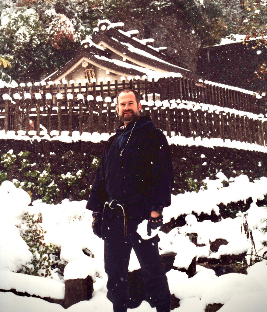 A young Stephen K. Hayes with brown hair and beard wears a black martial arts uniform and jacket while standing in front of a shrine in the deep, falling snow of Togakure Mountain.