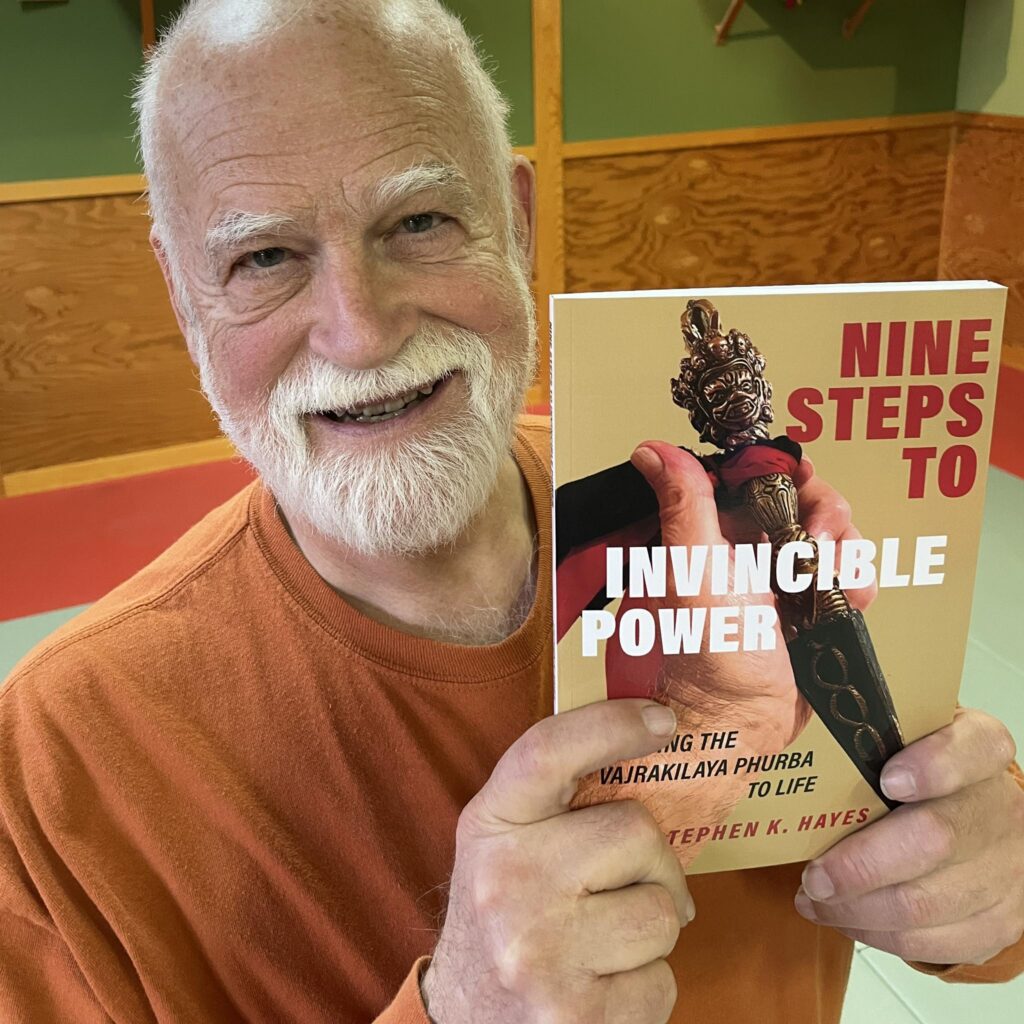 Stephen K. Hayes stands in his home dojo, smiling as he holds up a copy of his new book: Nine Steps to Invincible Power. The cover of the book features a hand holding a Tibetan phurba dagger.